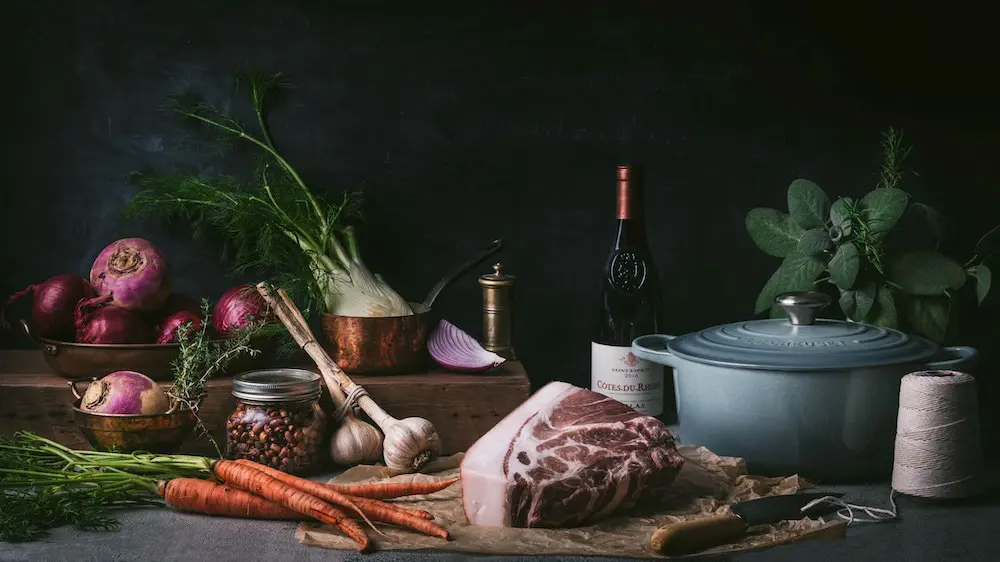 An arrangement of vegetables, a cut of beef, a bottle of wine and a Dutch Oven on a table