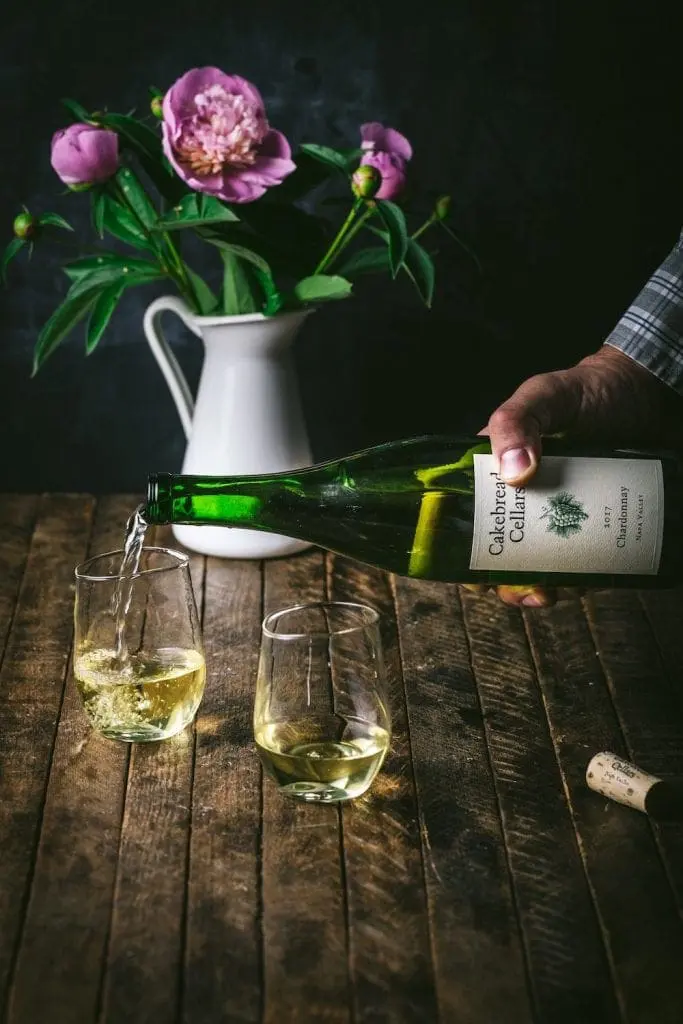 Pouring two glasses of white wine, with pink peonies in the background