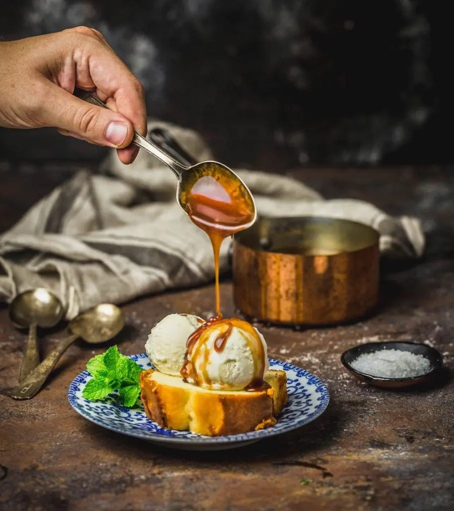 A hand pouring caramel from a spoon onto a bowl of ice cream and cake