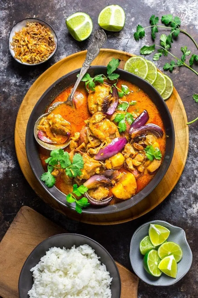 A bowl of colorful red curry with rice and limes around it