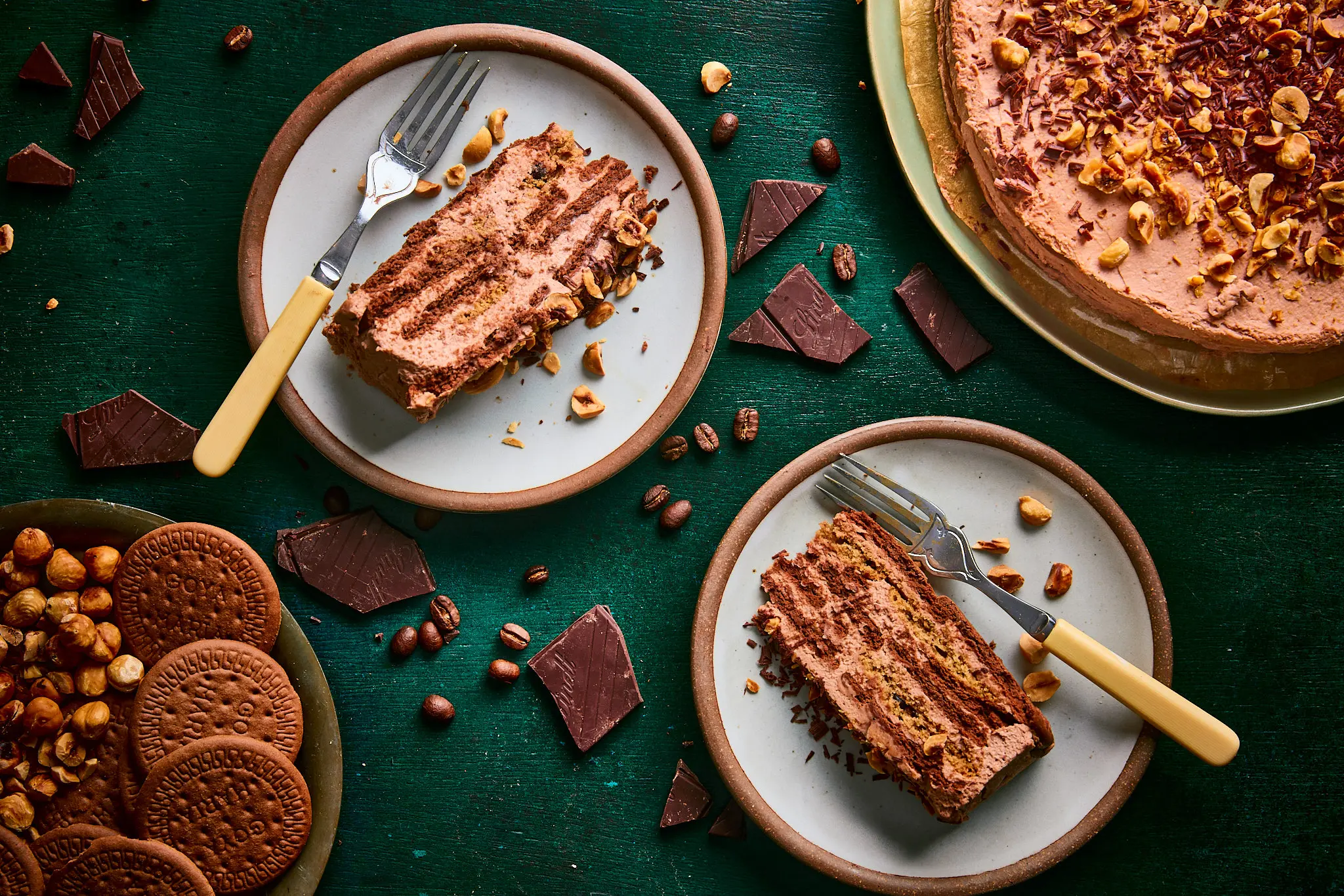 Two slices of icebox cake with a platter of cookies, chocolate, and hazelnuts.