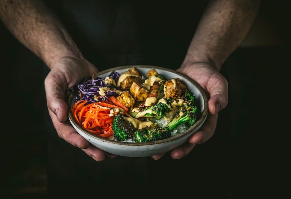 Hands holding a bowl with tofu, veggies and drizzled sauce
