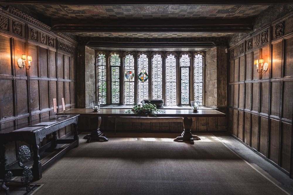 The wood-paneled interior of a Tudor mansion with stained glass windows in the background