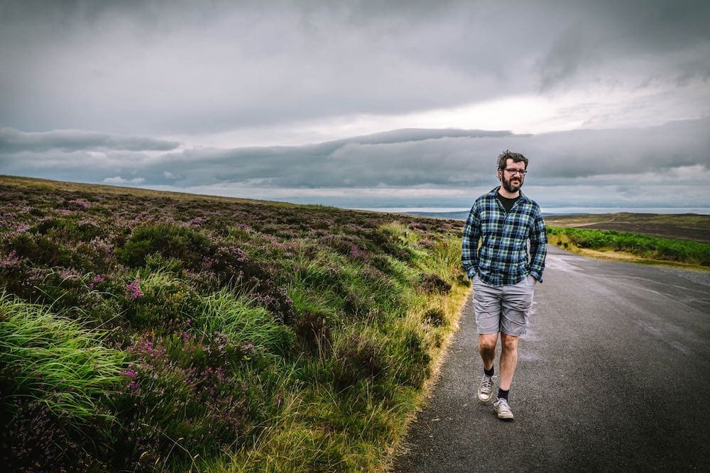 A wide, low moor with purple heather and a man walking towards camera