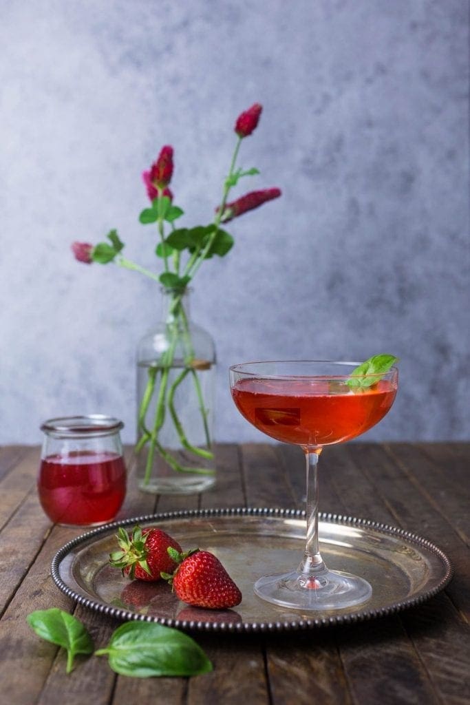 A bright red cocktail with strawberries on a plate and red flowers in a vase