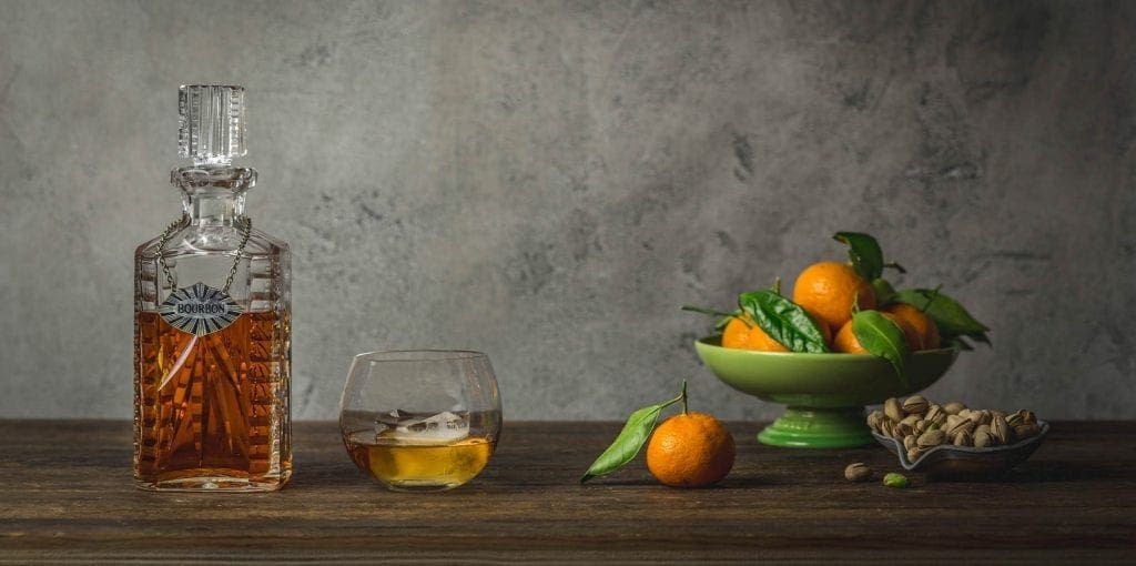 A decanter of whisky with a glass of whisky and a bowl of small tangerines